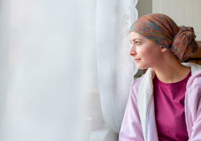 Young adult female cancer patient wearing headscarf and bathrobe sitting in the kitchen looking out window.