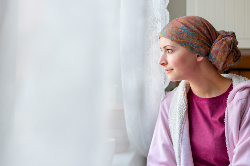 Young adult female cancer patient wearing headscarf and bathrobe sitting in the kitchen looking out window.